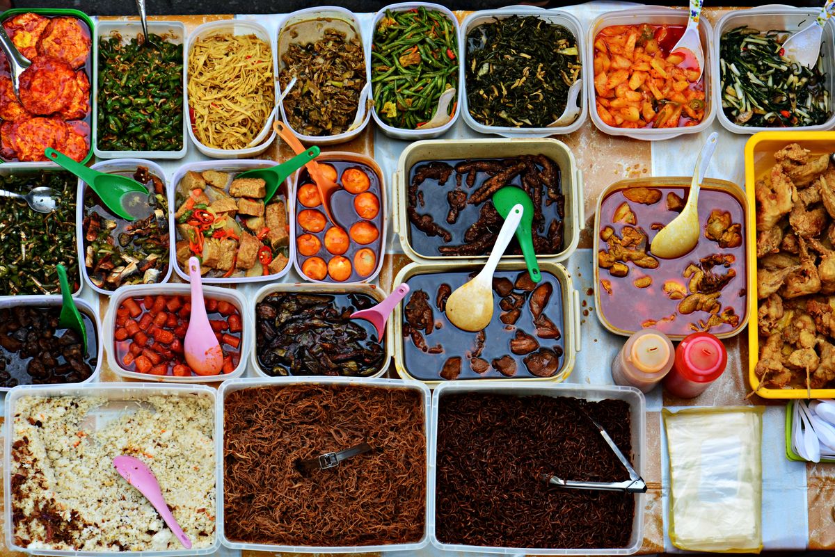 Overhead view of a mixed-rice stall counter, trays of curries, greens, fried chicken and serunding with serving spoons