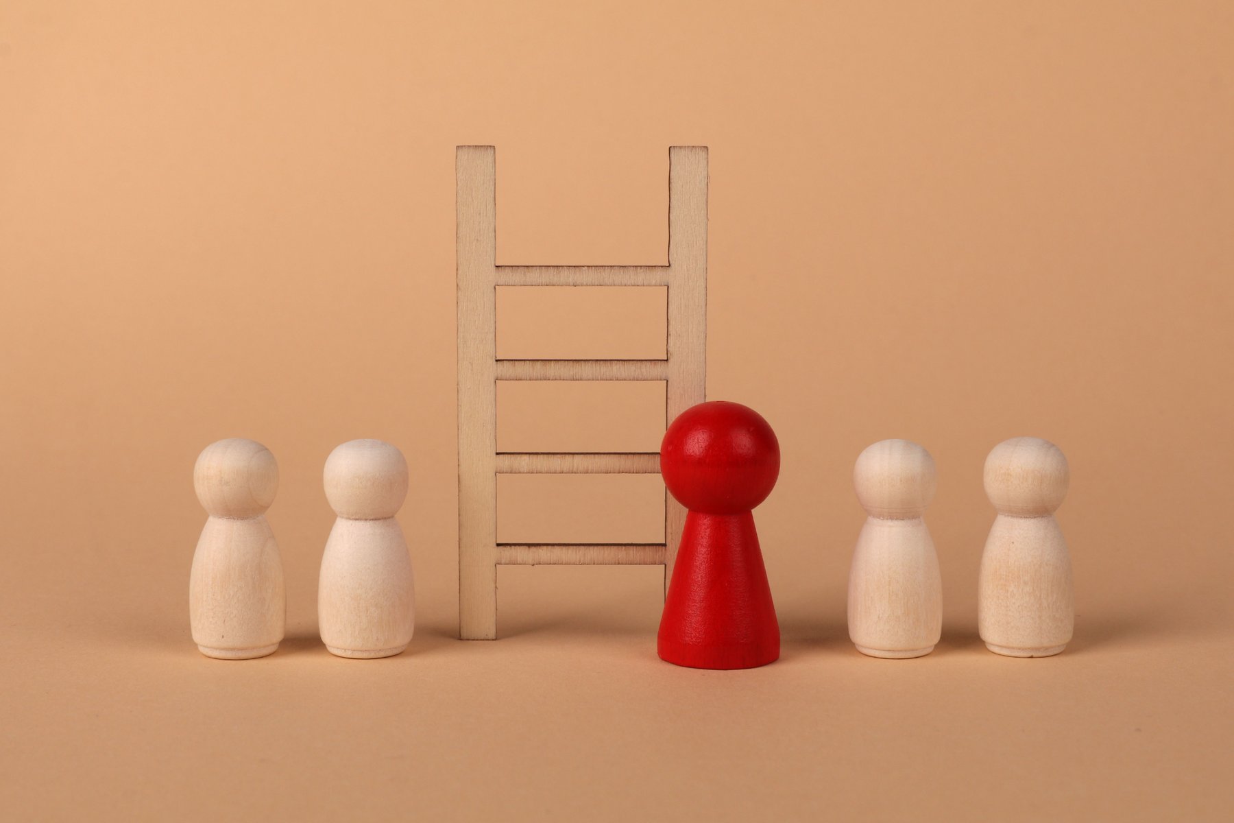 Five plain wooden peg figures in a line on a beige background, one of them red, beside a small wooden ladder
