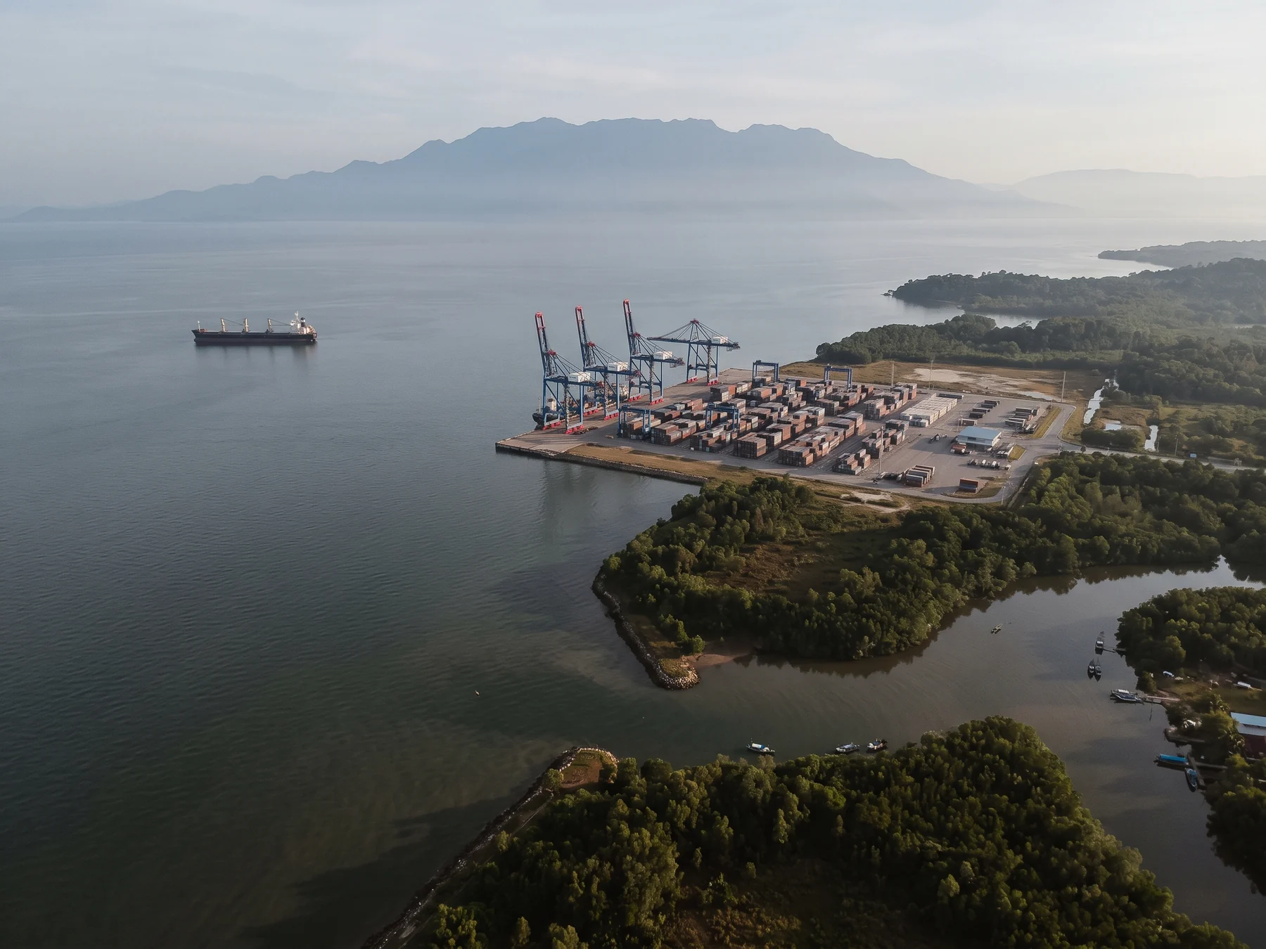 Aerial view of a tropical coastline at dawn, with a container terminal and gantry cranes on a reclaimed spit, a bulk carrier at anchor offshore, mangrove-fringed river mouths in the foreground and a long mountain range in silhouette through the haze