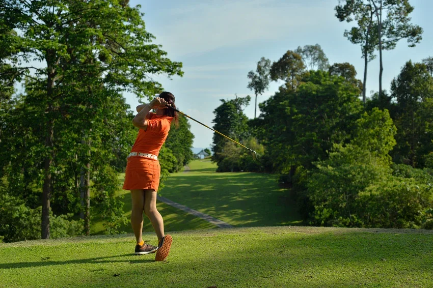 A golfer in orange at the end of her swing on a tee, fairway and rainforest behind her